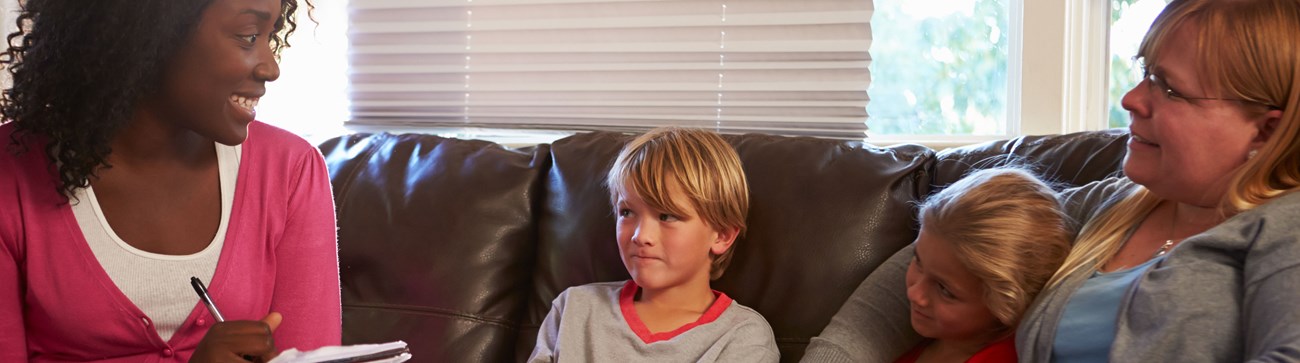 woman smiling at two young children whilst taking notes, one child under another womans arm, all sat on brown leather sofa