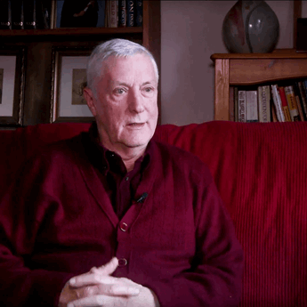 a man sat on a red sofa with his hands crossed, in the middle of talking