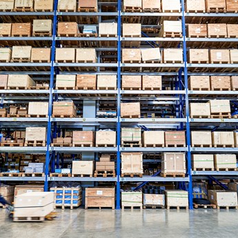 tall factory shelves filled with boxes, workman walking across wearing hard hat and overalls