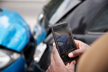 close up of hands holding mobile phone taking a photo of a blue car and a black car which have collided